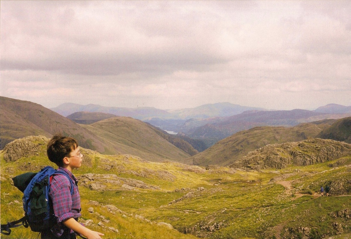 A boy in glasses and a plaid shirt, rucksack on, gazing across the rolling green fells of the Lake District under a heavy grey sky.